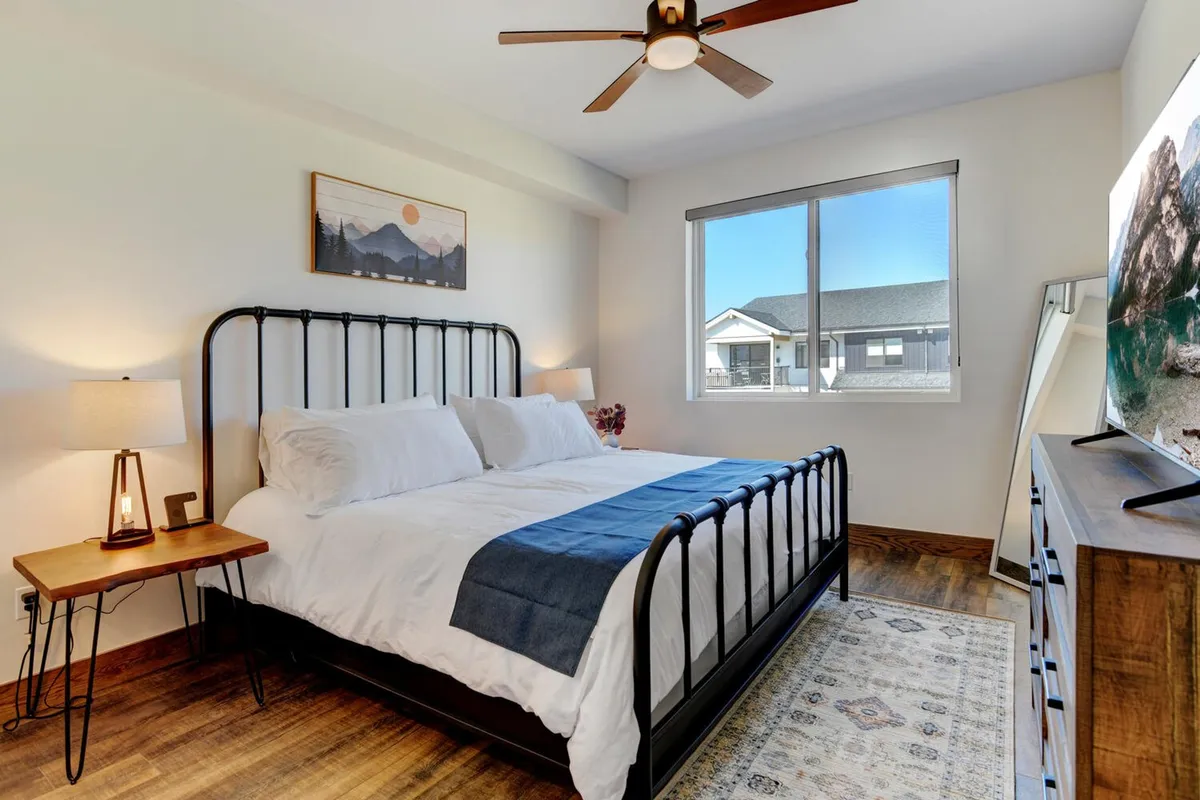 This image showcases a bedroom with white linens and a blue throw blanket, a wooden nightstand with a lamp, a large window letting in natural light, and decorative wall art. The room feels cozy and peaceful, with sunlight streaming in to enhance the relaxing atmosphere.