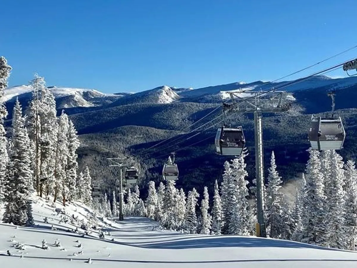 A panoramic view of ski lift gondolas moving above snowy slopes, with snow-covered trees and a breathtaking mountain range in the background. Skiers and snowboarders can be seen descending the slopes, highlighting the active winter ambiance.