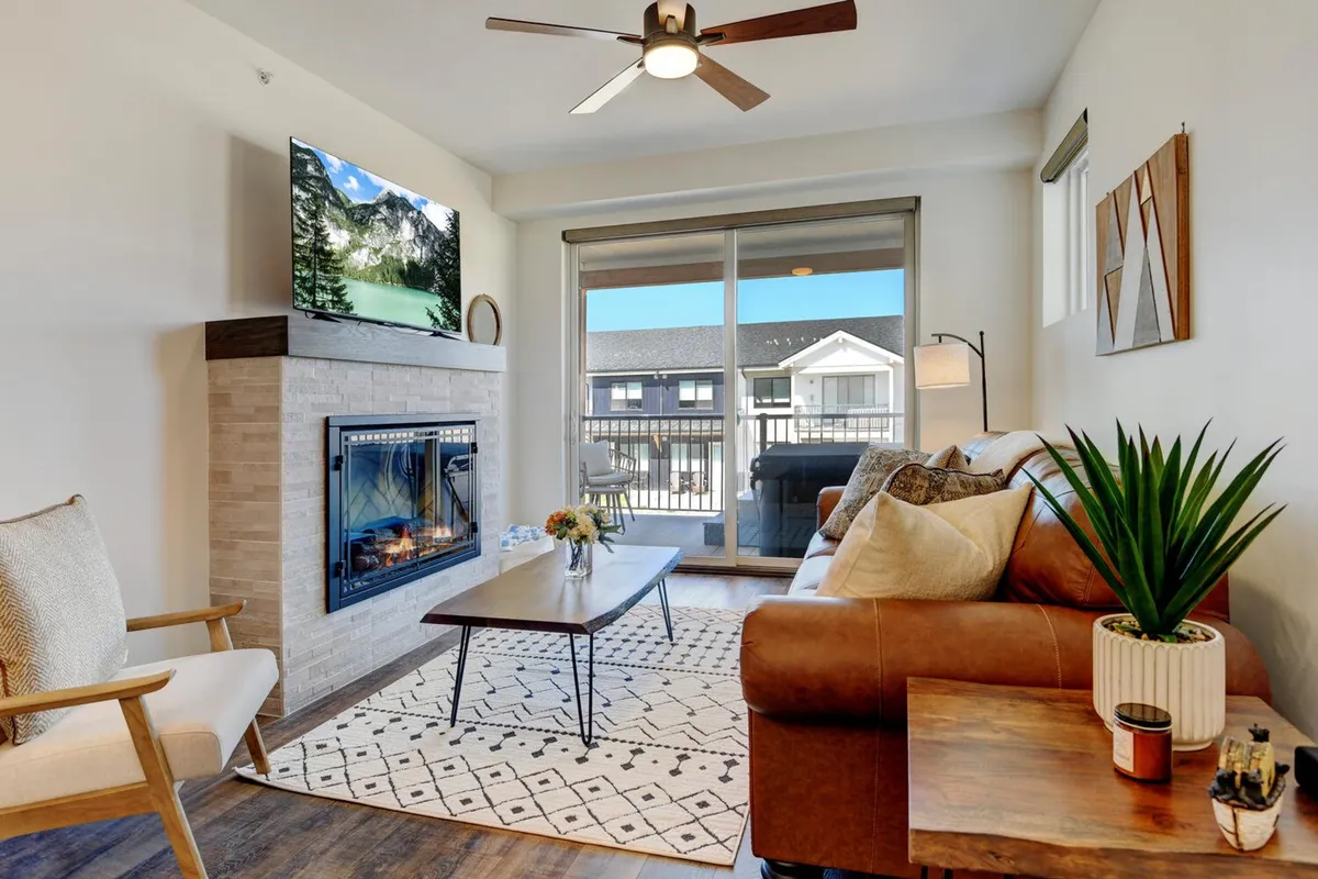 The image showcases a cozy living room with a stylish fireplace as the centerpiece, surrounded by a comfortable sofa, coffee table, and lush green plants. Natural light pours in through large windows, illuminating the inviting space that’s perfect for gathering, relaxing, and enjoying entertainment on the mounted television.
