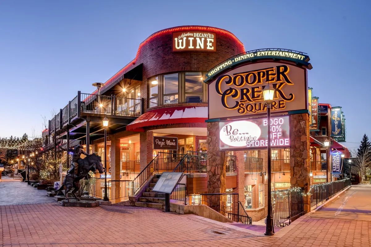 A lively downtown scene featuring the Cooper Creek Square sign, a wine shop, outdoor seating, and a sculpture, with people gathering to shop, dine, and enjoy live entertainment.