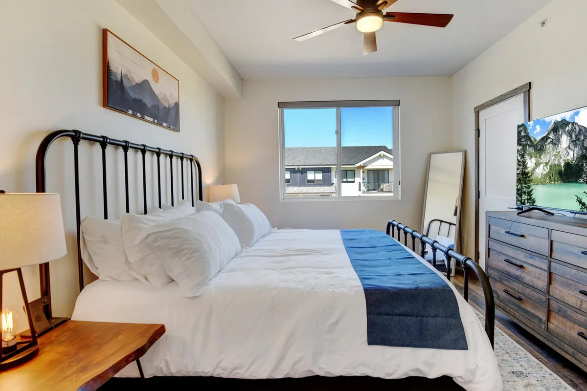 A serene bedroom with crisp white linens, a blue throw, soft natural light, and thoughtful décor like mountain artwork and a side table. The room radiates tranquility and suggests premium comfort with luxury linens and a calming retreat vibe.