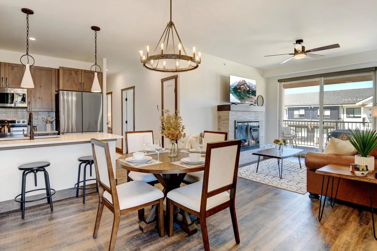 This interior image features a cohesive view of an open-concept gathering space, including a welcoming living area with a plush sofa, a modern electric fireplace, a dining table, and a visible kitchen island. The mood is cozy and inviting, with warm lighting and tasteful decor that conveys both comfort and style. The fireplace and sofa invite relaxation, while the dining table and kitchen island suggest shared meals and lively conversations.