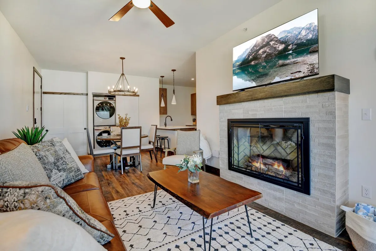 A warm, welcoming interior scene featuring a stylish fireplace with a coffee table and a comfortable sofa in the foreground. The dining area is visible further back, and a large TV is mounted above the fireplace. The arrangement is ideal for relaxing, entertaining, or watching movies, reflecting a blend of mountain charm and modern amenities.