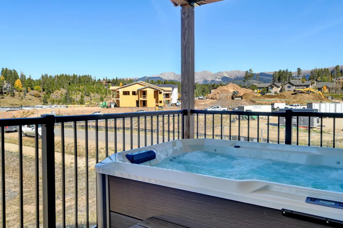An inviting outdoor hot tub set against a stunning mountain backdrop at sunset. The scene includes groups of people relaxing in the hot tub, surrounded by trees and panoramic views, evoking a sense of relaxation and connection to nature. The presence of the hot tub and mountain setting hints at resort-like amenities and an exclusive, tranquil atmosphere.